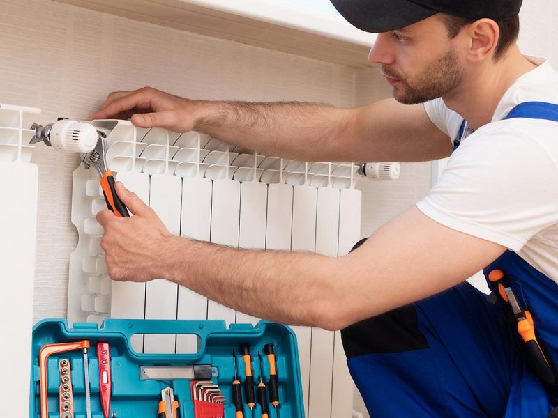 Professional young repairman in special uniform with tools is installing radiators and thermostat in domestic room