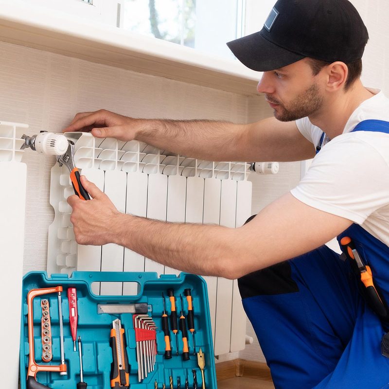 Professional young repairman in special uniform with tools is installing radiators and thermostat in domestic room